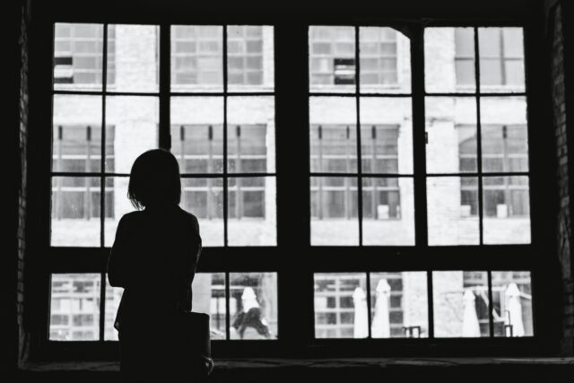 Silhouette of woman standing inside wall-length windows looking out on street - building visible across the street.